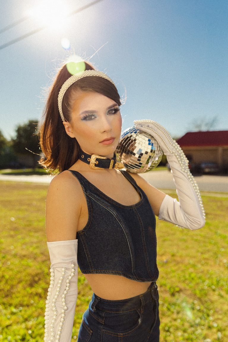 Gabriella Stella press photo outside wearing white gloves along with a fashion belt wrapped around her neck