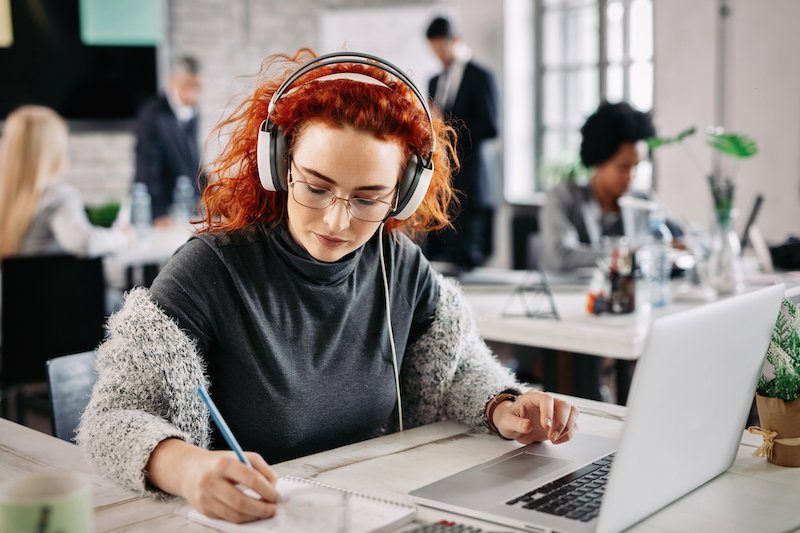 Redhead businesswoman using laptop and writing notes in her notepad while listening music on headphones at work.