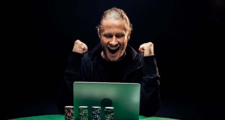 happy man gesturing while using laptop near poker chips isolated on black.